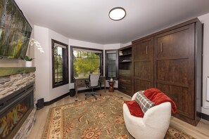 A room with a fireplace, a large window with a view of trees, a desk with a chair, and a large wooden cabinet. - New 5 Bedroom Luxury Mountain Retreat w/Hot Tub, Private Wooded Lot - Sleeps 8 (Silverthorne)