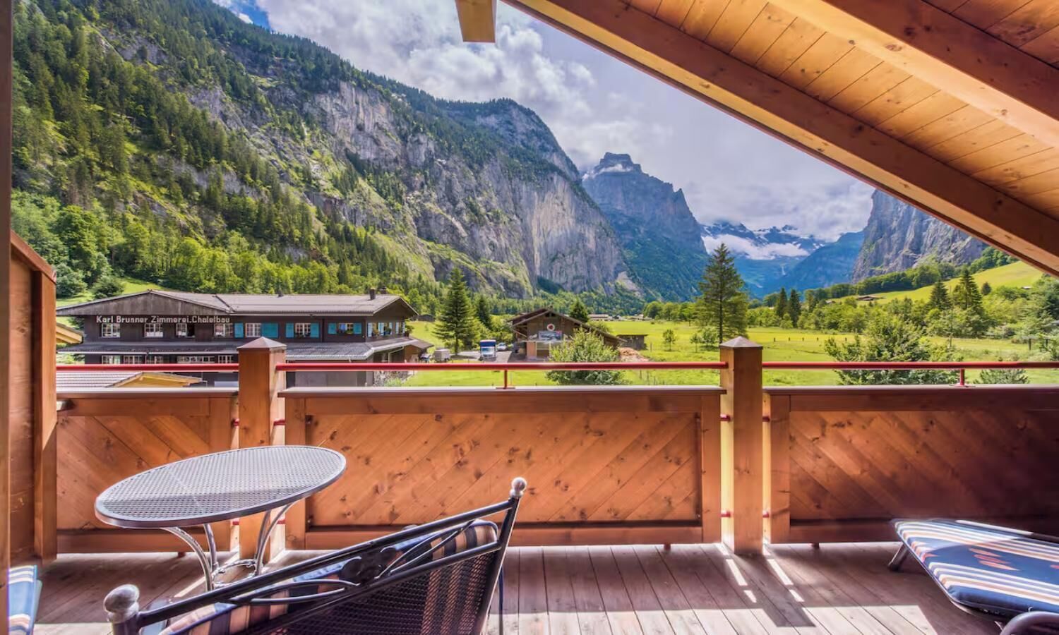 A wooden deck with a table and chairs, overlooking a mountainous landscape with a building labeled 'Karl Brunner Zimmerle Chaletsbau'.
