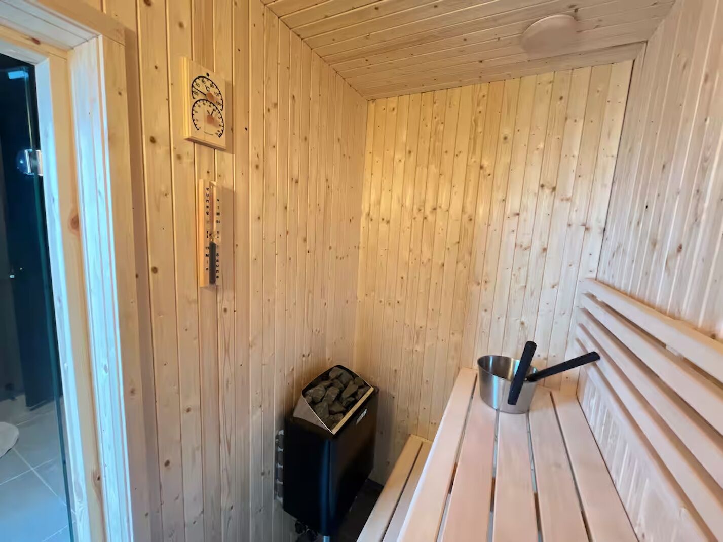 A wooden cabin interior with a wooden bench, a metal bucket, a small heater, and a wooden door.