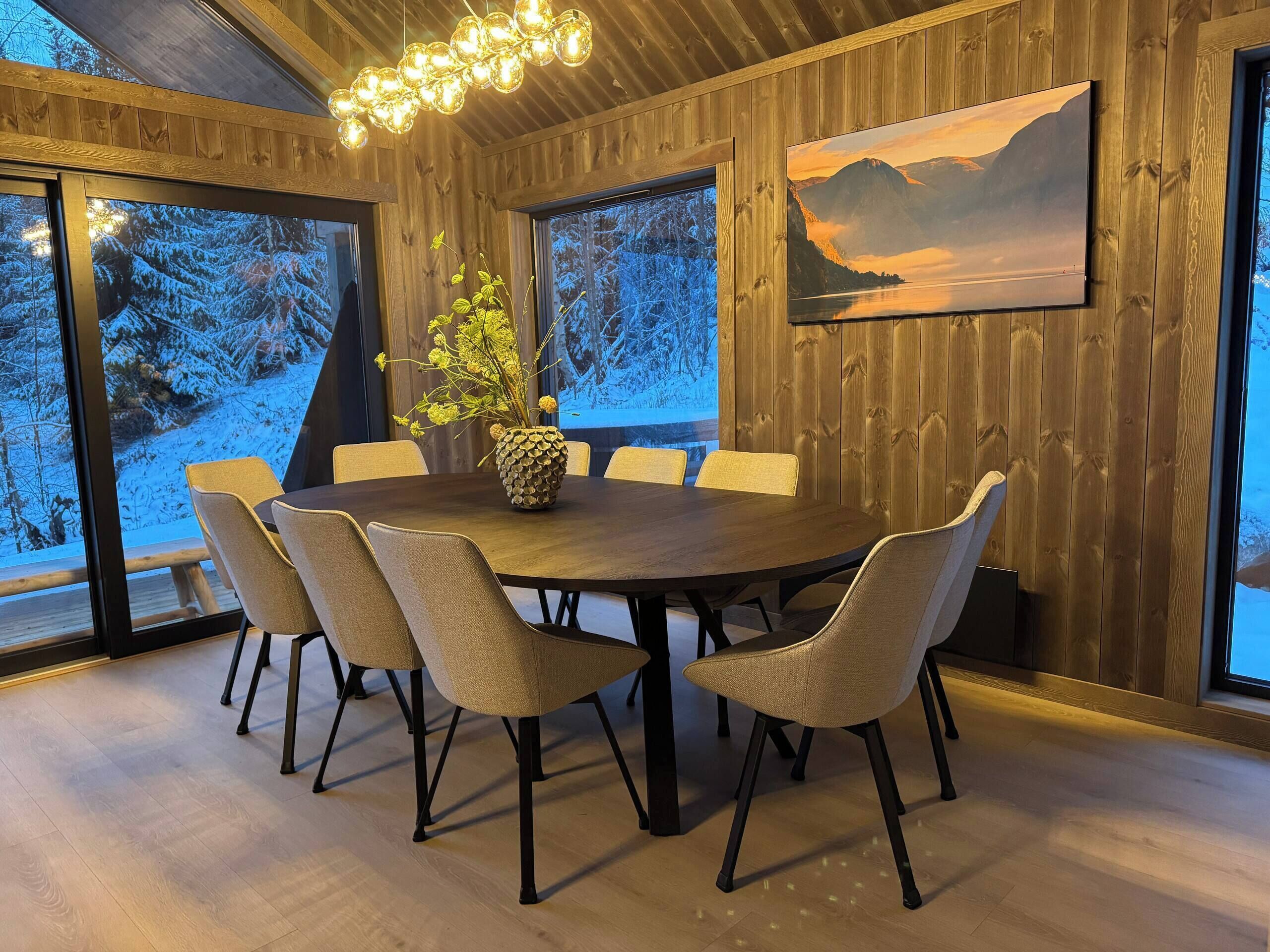 A dining area with a round table, six chairs, a vase with flowers, wooden walls, and a painting of a mountain landscape.