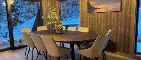 A dining area with a round table, six chairs, a vase with flowers, wooden walls, and a painting of a mountain landscape.