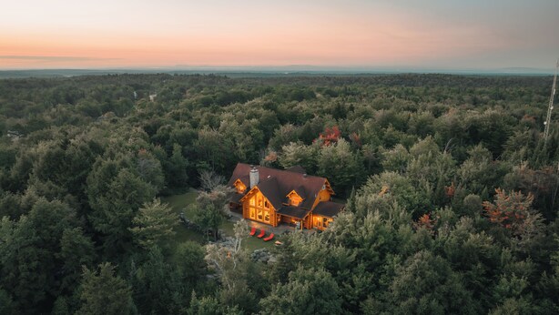 A large log cabin surrounded by dense forest, with a clear view of the surrounding landscape. - LUXURY LOG CABIN (Mille-Isles)