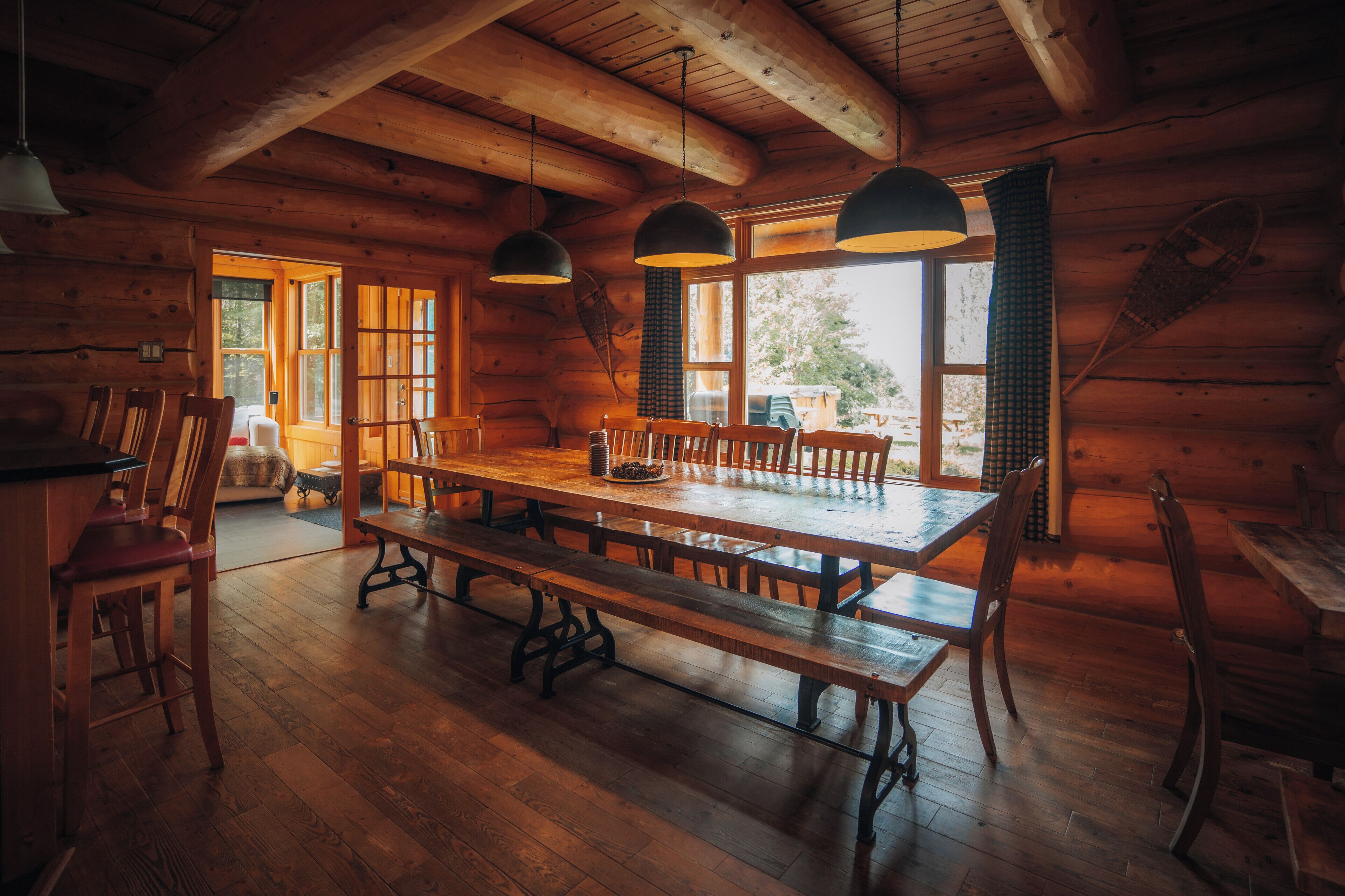 Ein Holzblockhaus-Interieur mit einem großen Esstisch, Bänken und hängenden Pendelleuchten.
