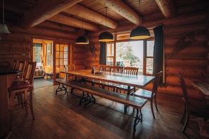Interior de una cabaña de madera con una gran mesa de comedor, bancos y lámparas colgantes.