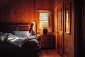 Una habitación de cabaña de madera con una cama, una mesita de noche con una lámpara y un ventanal.