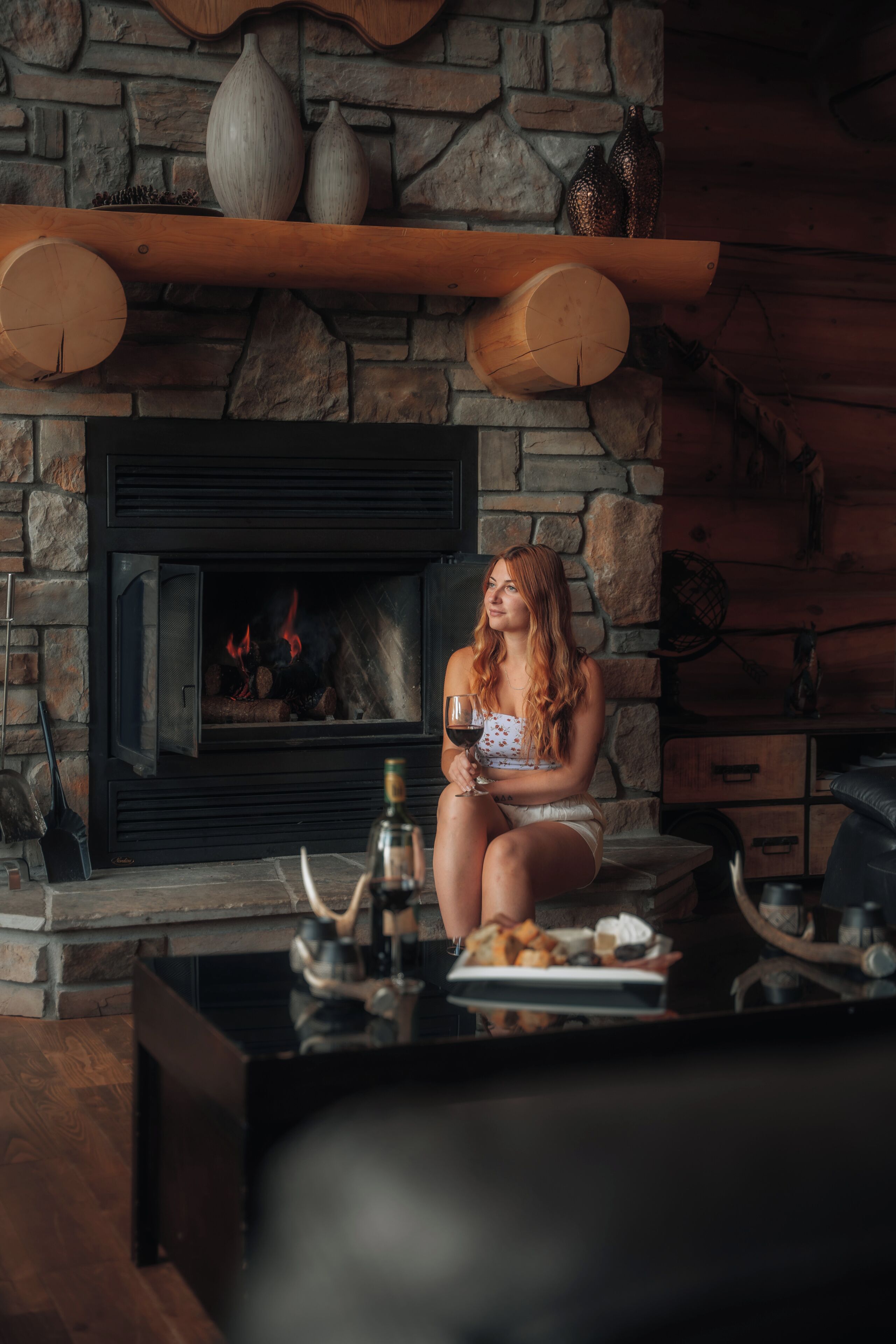 A woman sitting by a fireplace with a glass of wine, surrounded by stone walls and wooden furniture.