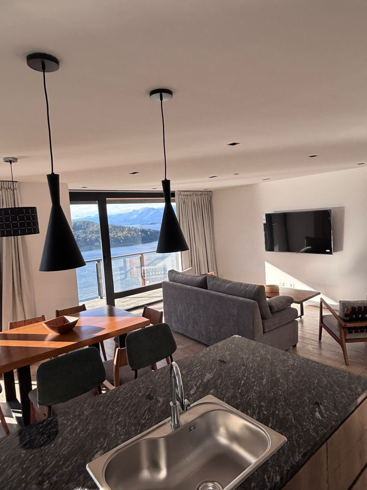 Une cuisine moderne dotée d’un évier encastré, d’une table à manger et de chaises. Une grande fenêtre offre une vue sur les montagnes et un plan d’eau.