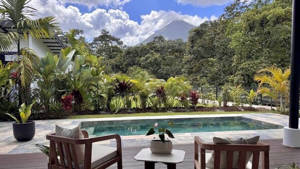 Un espace aménagé au bord de la piscine, avec deux chaises en bois et une table, entouré d’une végétation tropicale luxuriante et offrant une vue sur les montagnes.