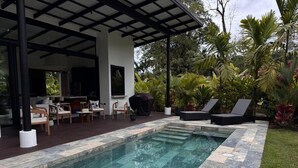 Une piscine rectangulaire bordée de pierres, entourée de chaises longues et d’un espace de détente couvert, comprenant des chaises et une table.