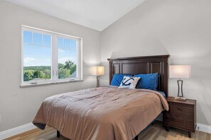 A bedroom with a large window, a bed with a brown bedspread, two bedside lamps, and a nightstand. - Ellison Bay 3BR Vacation Home - Water Views, Beach & Boat Parking (Ellison Bay)