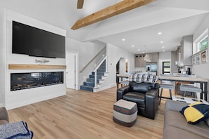 A modern living room with a flat-screen TV, a fireplace, a staircase, a dining area, and a kitchen. - Ellison Bay 3BR Vacation Home - Water Views, Beach & Boat Parking (Ellison Bay)