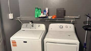 A laundry area with two front-loading washing machines, a shelf with cleaning supplies, and a hose.