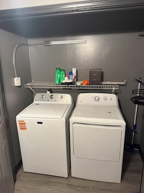 A laundry area with two front-loading washing machines, a shelf with cleaning supplies, and a hose.