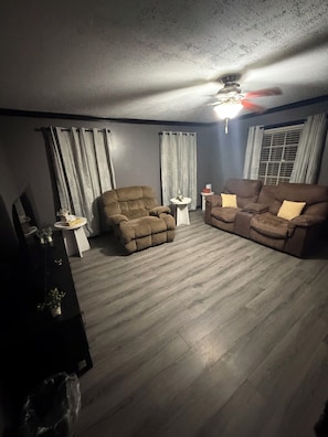 Un salon avec un ventilateur de plafond, un canapé marron, un fauteuil marron et une petite table blanche.