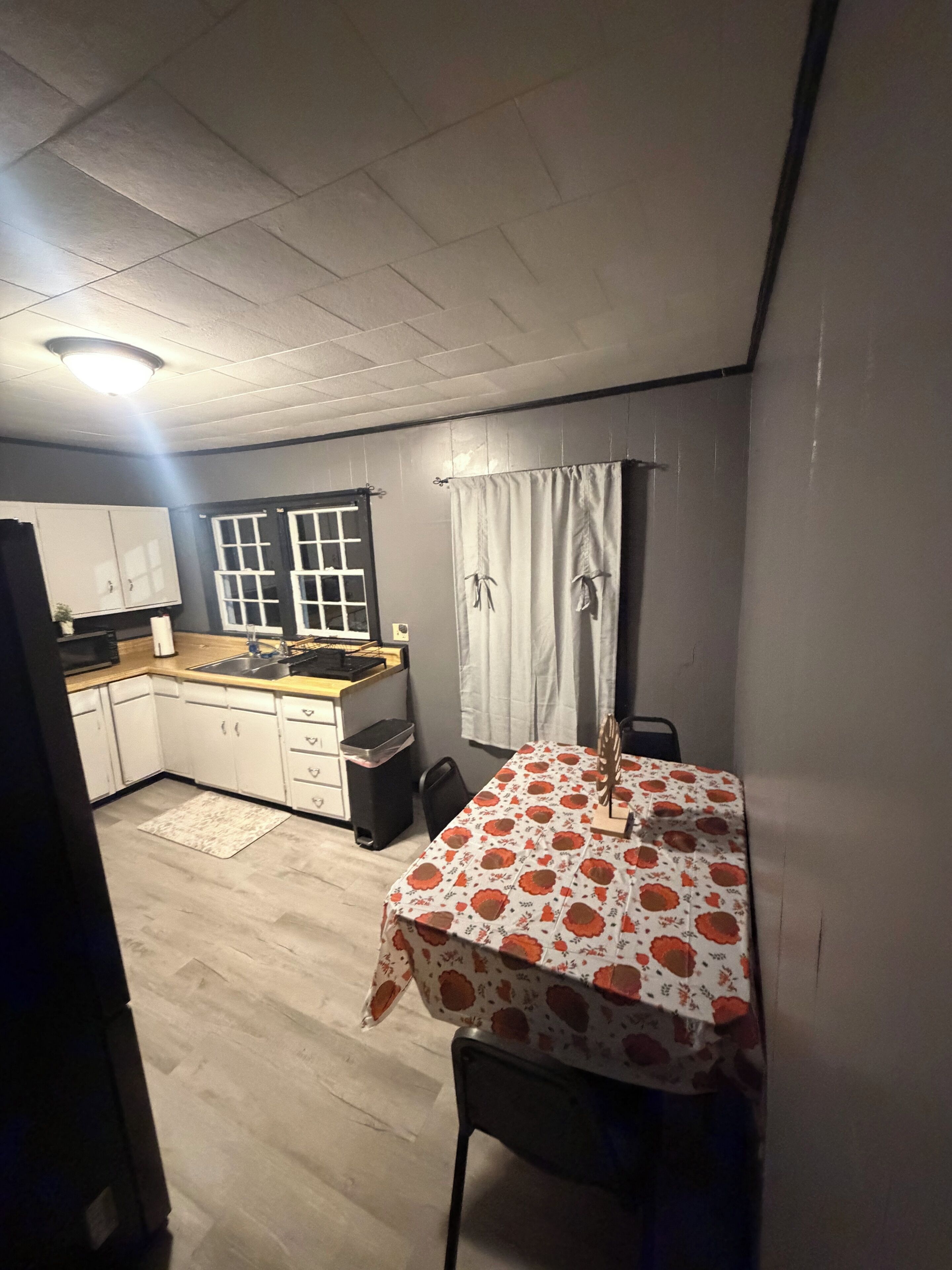 A compact kitchen with white cabinets, a microwave, and a sink. There's a dining table with a floral tablecloth.