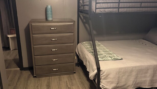 Quarto com uma cama de solteiro, cômoda com gavetas e uma pequena mesa com porta-retrato.