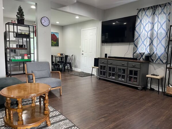 A living room with a wooden coffee table, a grey armchair, a dining area with a table and chairs, a kitchen area with a fridge and shelves, a clock on the wall, and a TV mounted on the wall. - 4-bedroom house in tranquil Baltimore Highlands with AC (Baltimore Highlands)
