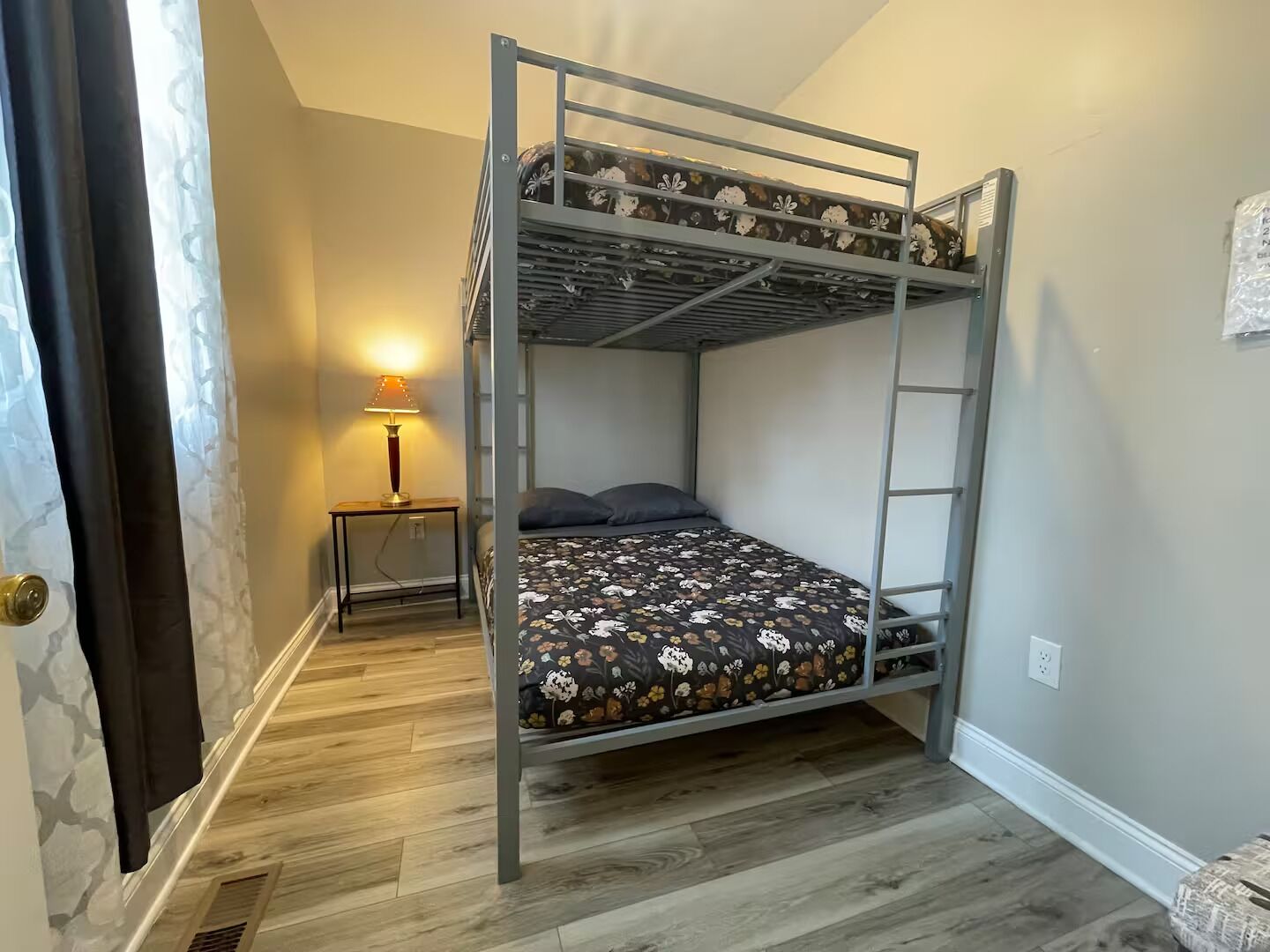 Une chambre avec un lit superposé, un plancher en bois, une table de chevet avec une lampe et une prise électrique au mur.