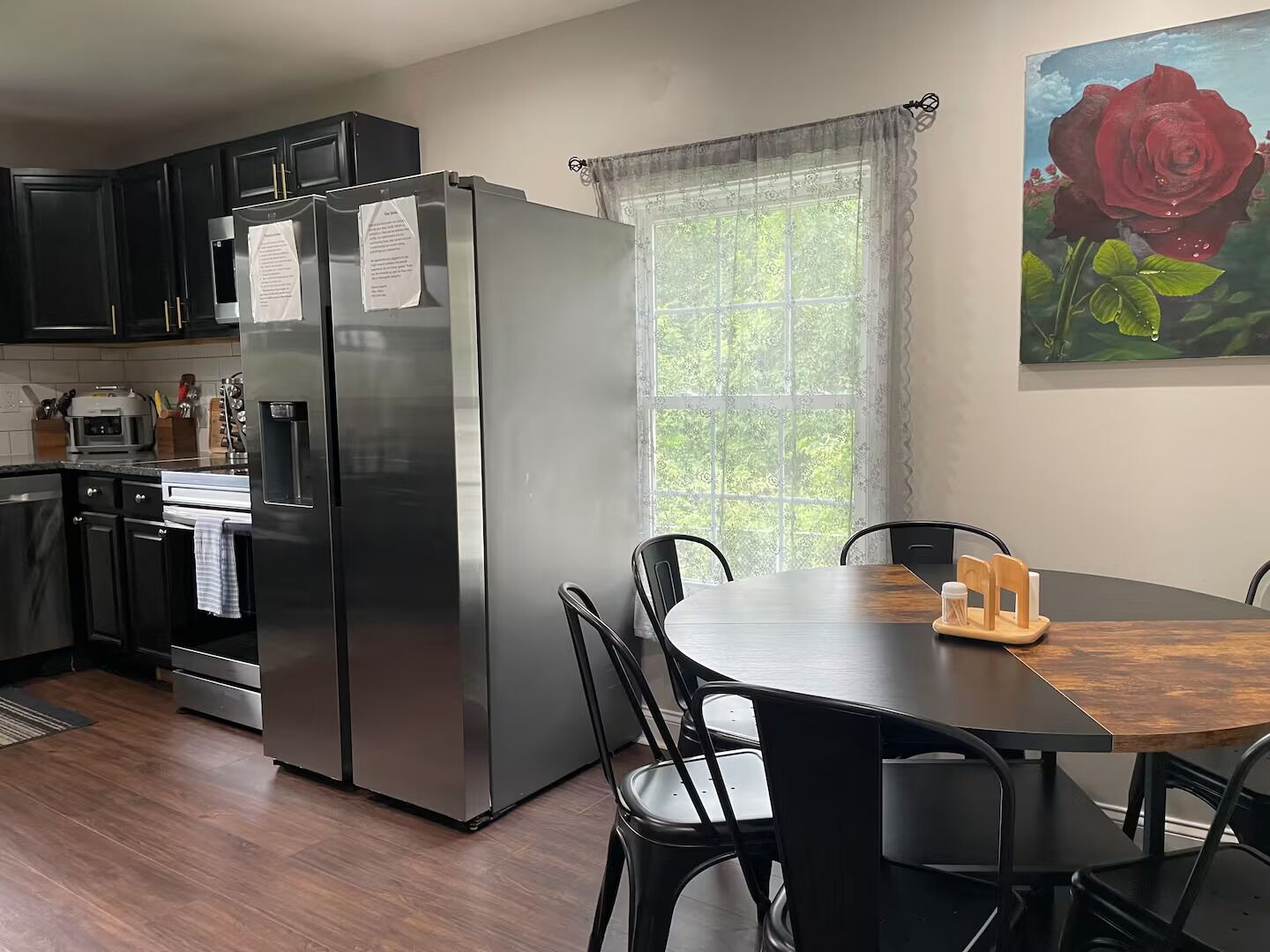 A modern kitchen with a stainless steel refrigerator, a dining table with chairs, and a window with a view of greenery.