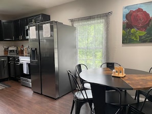 A modern kitchen with a stainless steel refrigerator, a dining table with chairs, and a window with a view of greenery.
