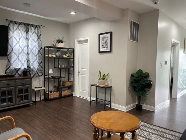 A living room with a wooden floor, a round table, a TV, a bookshelf, and a door.