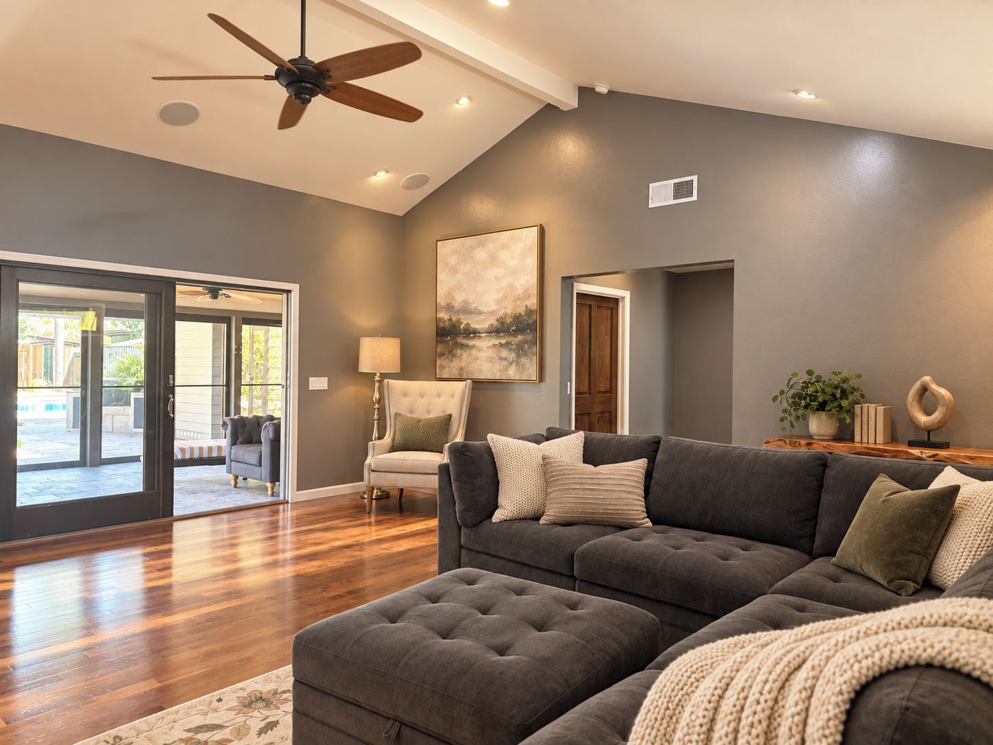 A spacious living room with a dark grey couch, a tufted ottoman, and a ceiling fan.