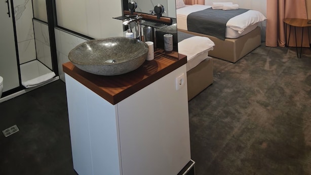 Una habitación de hotel moderna con un espejo grande, un lavabo redondo, una cama con colcha oscura y una pequeña mesa con una silla.