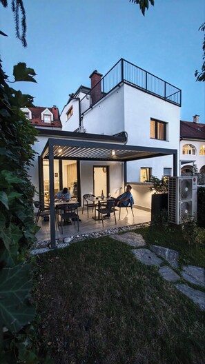 A modern outdoor patio with a pergola, seating, and a person working at a table. - Huch House Hotel and apartments (Kranj)