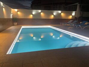 Una piscina rectangular con borde de azulejos y un borde iluminado, rodeada por un muro bajo con luces uniformemente espaciadas.