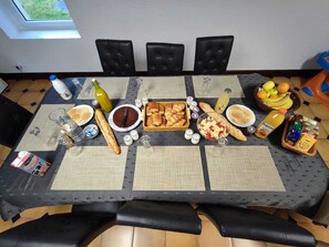 Ruokapöytä on katettu erilaisilla ruoilla ja juomilla, ja sen ympärillä on mustilla kankailla verhoiltuja tuoleja.