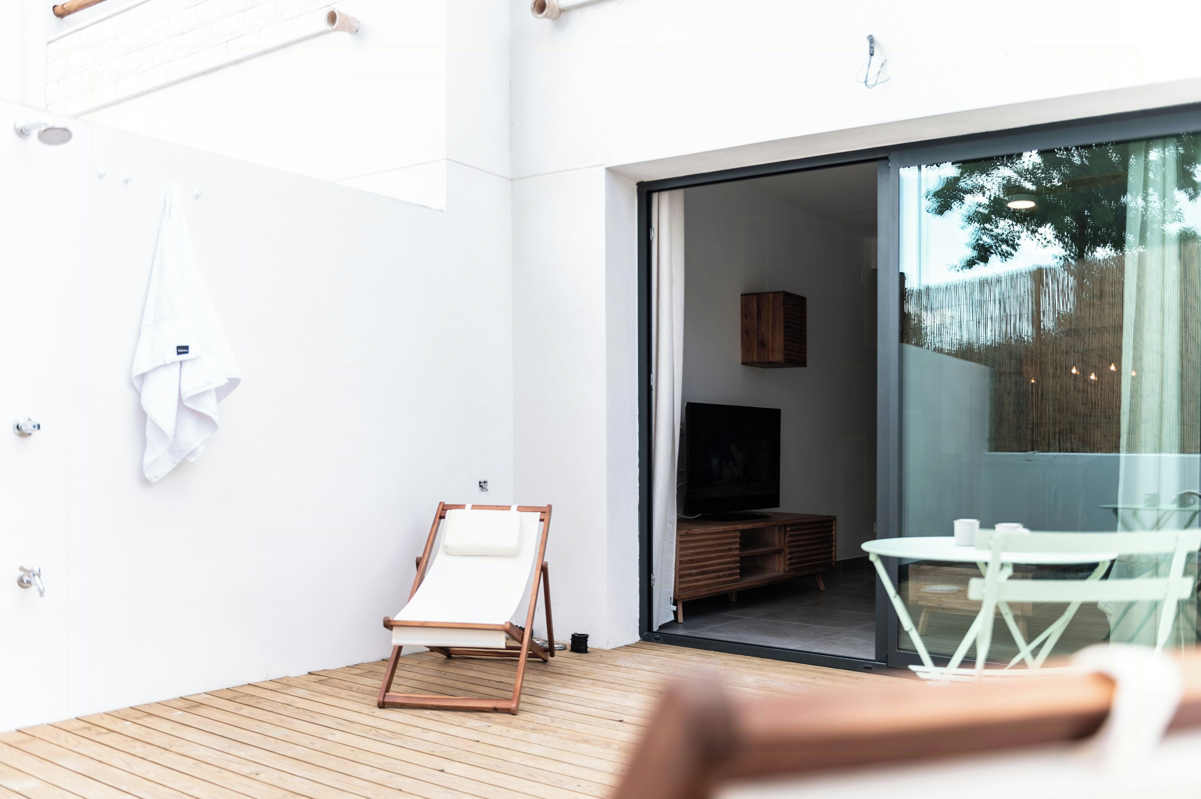 A modern outdoor space with a wooden deck, a lounge chair, a small table, and a sliding glass door leading to an interior room with a television.