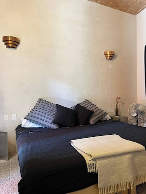 Un dormitorio con cama, almohadas, una mesita de noche, una lámpara y un aplique de pared.