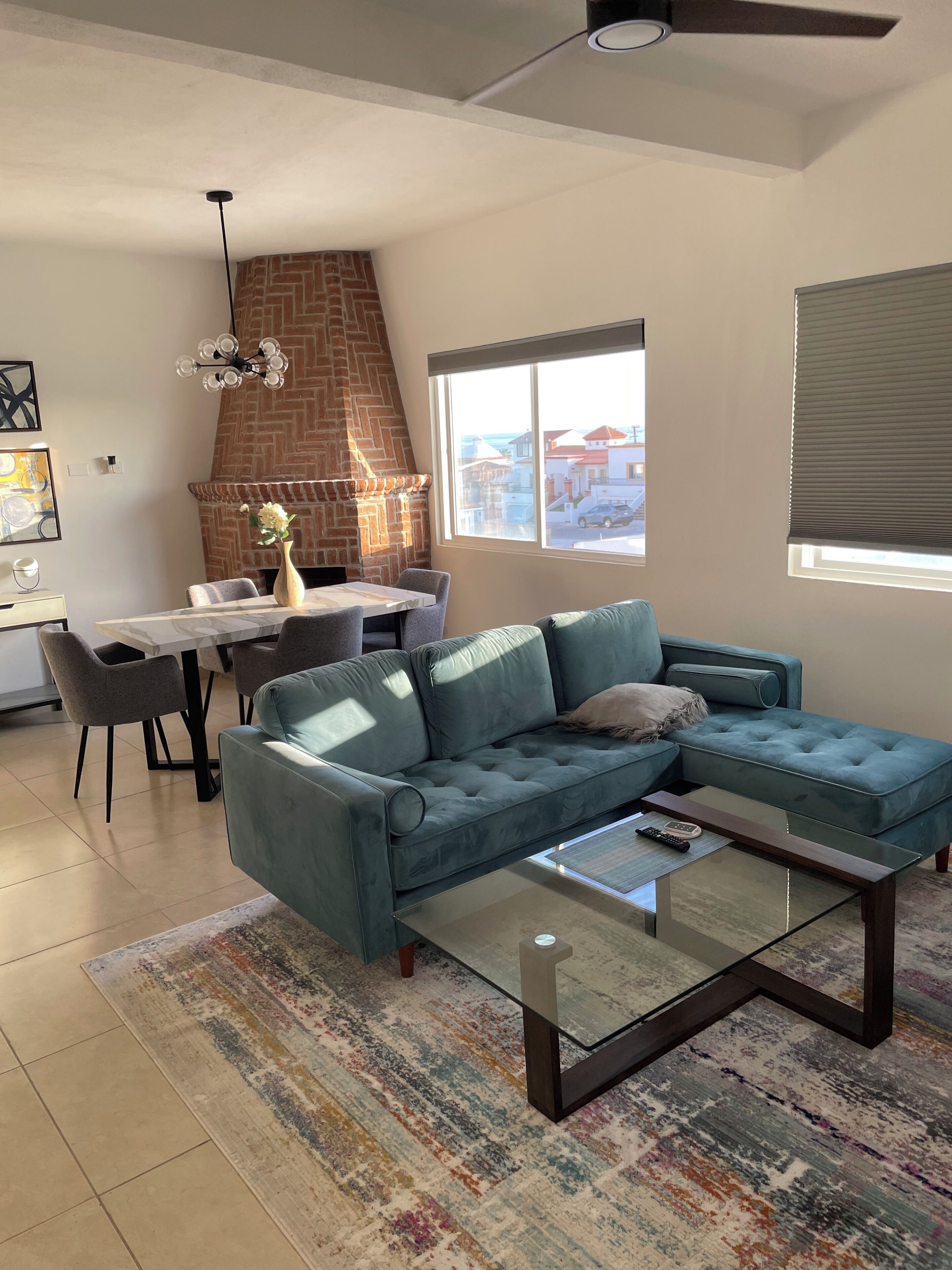 Een woonkamer met een hoekbank, een glazen salontafel, een eethoek met tafel en stoelen, een metselwerk open haard en een groot raam met zonwering.