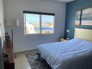 A bedroom with a bed, a nightstand, a lamp, a window with a view of buildings, and a decorative item on the nightstand. - Serene 2-bedroom condo in Rosarito perfect for relaxing getaways (Rosarito)