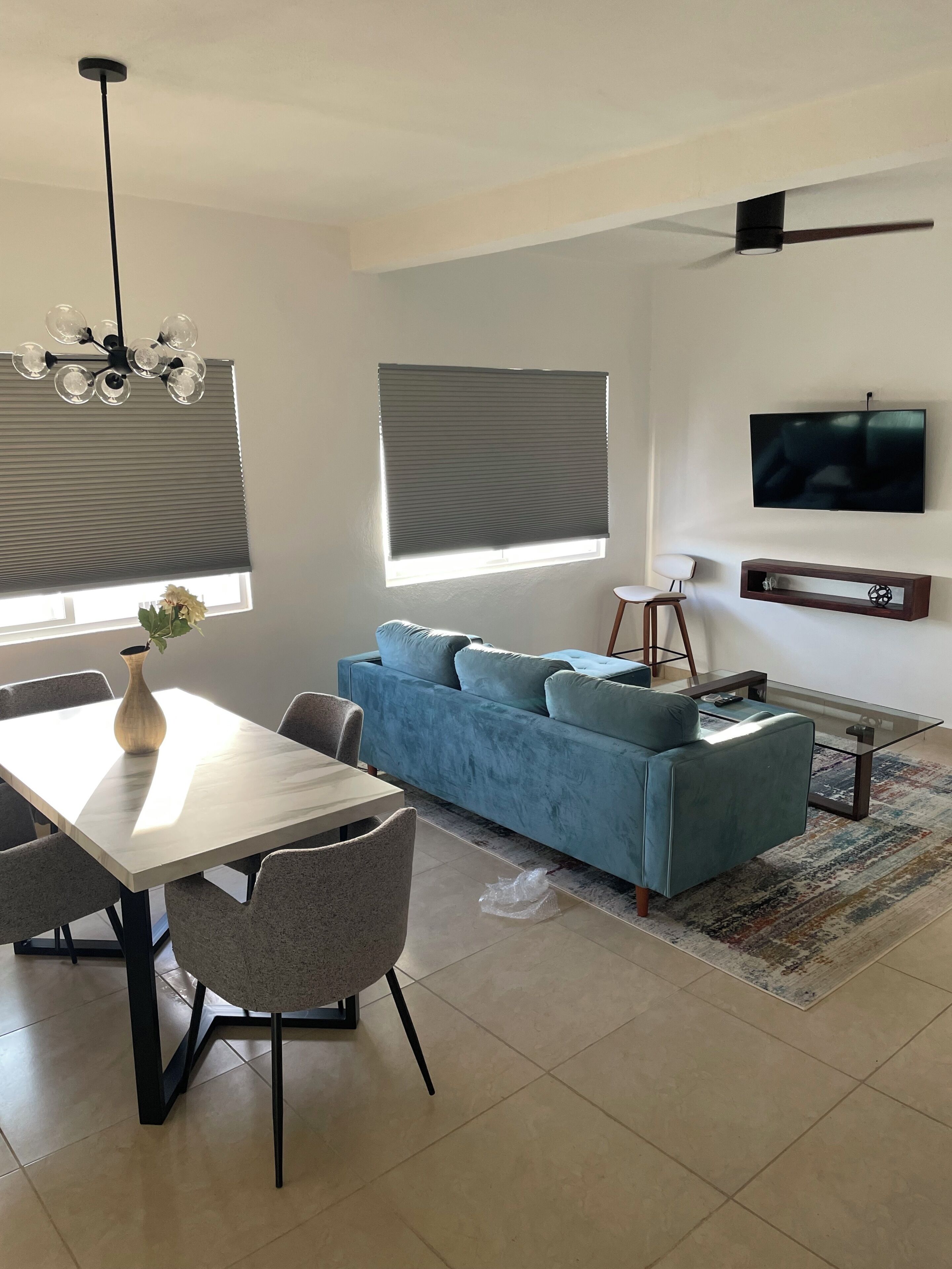 Una sala de estar moderna con un sofá, una mesa de comedor con sillas, un televisor montado en la pared y un ventilador de techo.