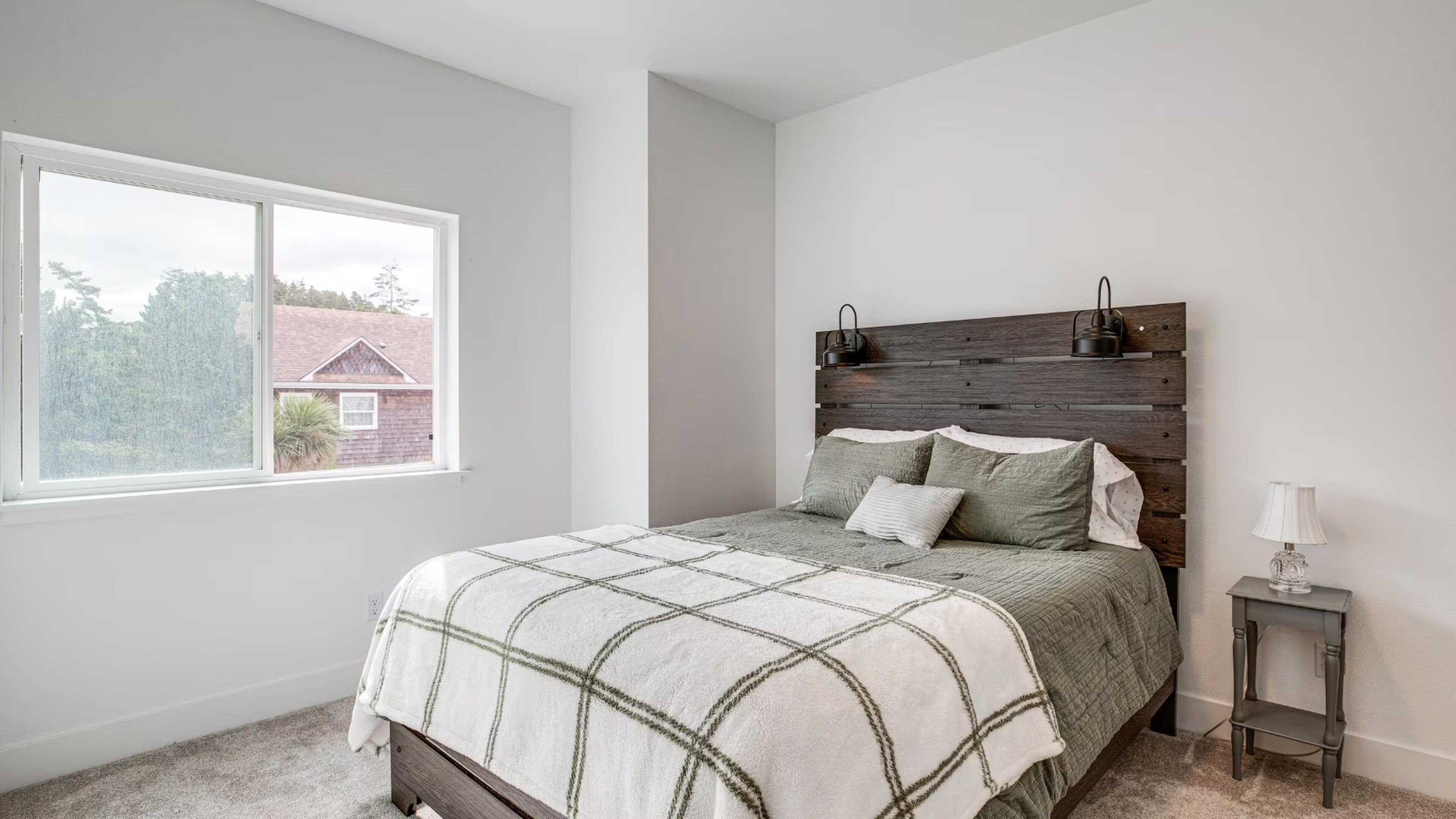 A bedroom with a bed, a nightstand, a lamp, and a window with a view of a house.