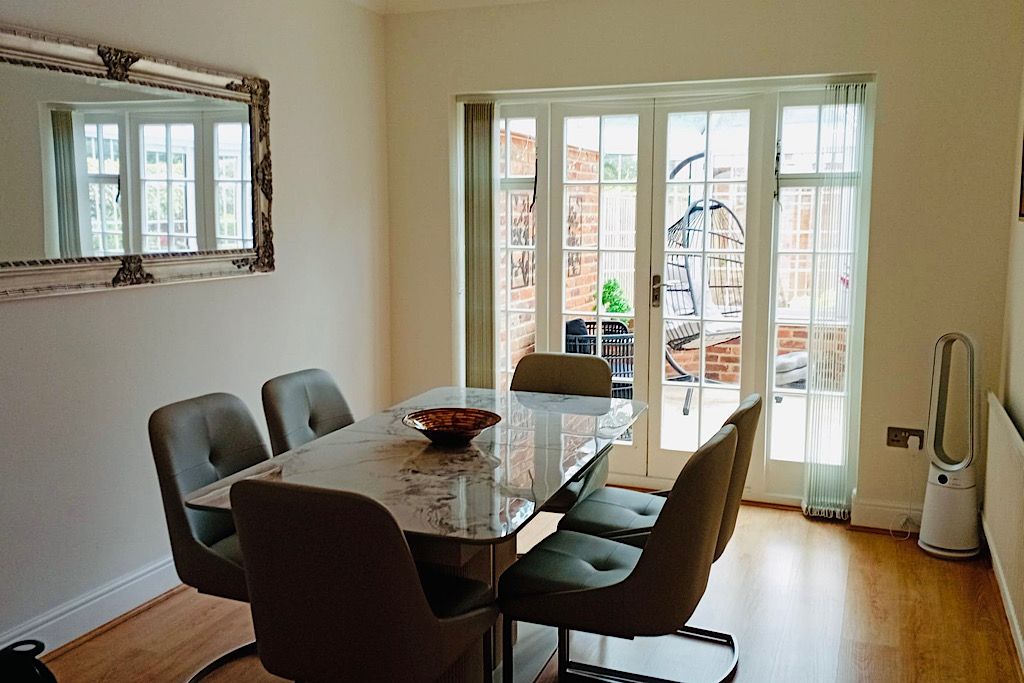 A dining area with a glass table, four chairs, a large mirror, and a view through a glass door to the outside.