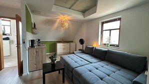 A modern living space with a grey sofa, a small table, a kitchen area with a fridge, and a window with a plant. - Maisonette-Wohnung am Markt  zentral in Wolkenstein, für 4 Gäste (Wolkenstein)