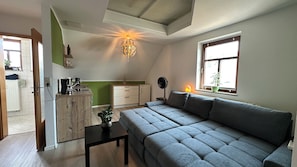 Ein stilvolles Wohnzimmer mit grauem Sofa, einem kleinen Tisch, einer Küchenzeile mit Kühlschrank und einem Fenster mit Pflanze.
