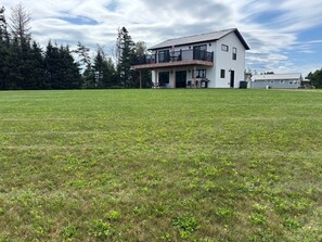 A two-story house with a balcony, surrounded by a grassy field and trees. - Rustic Bay Vacation Home (Lot 65)