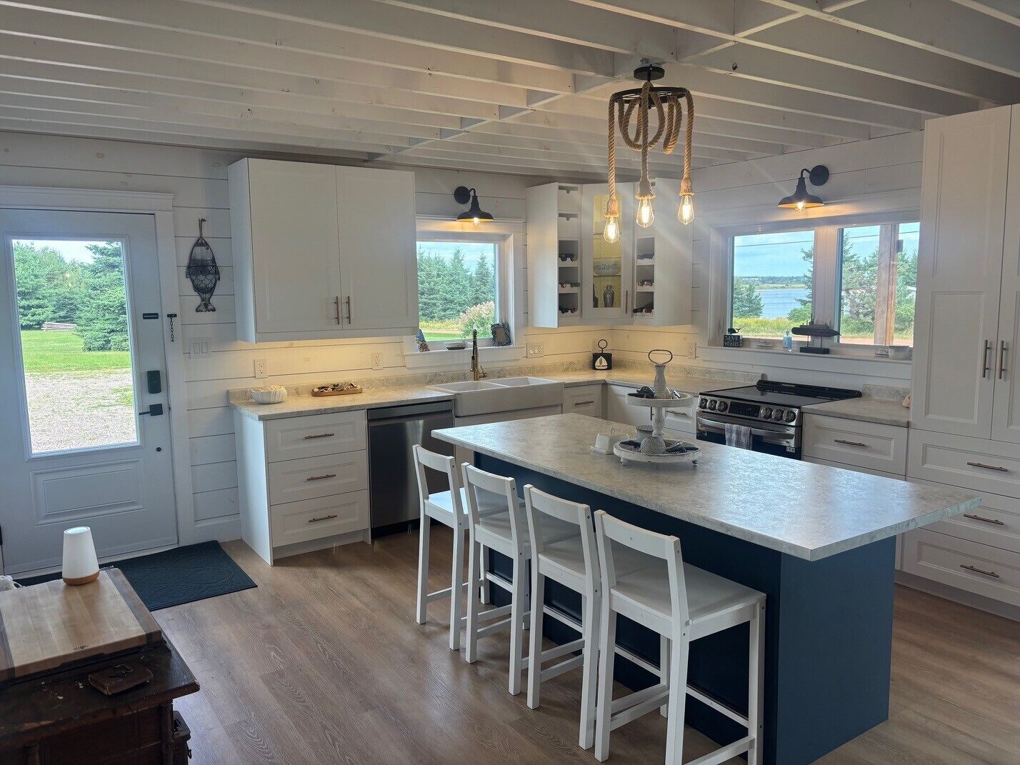 Une cuisine moderne avec des meubles blancs, un îlot central doté de tabourets de bar et une vue sur l’extérieur grâce à de grandes fenêtres.