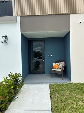A modern entrance with a glass door, a small outdoor seating area with a chair and a decorative pillow, and a pathway leading to the door. - Elegant and cozy apartment on Hoestead (Homestead)