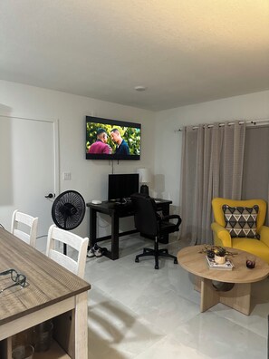 A living room with a television, a desk with a computer, a yellow armchair, a dining table with chairs, and a fan. - Elegant and cozy apartment on Hoestead (Homestead)