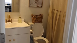 A bathroom with a toilet, a sink with a gold faucet, a wicker basket on the toilet tank, a framed picture on the wall, and a beige shower curtain. - Tranquil 1-bedroom cottage in the pines in Mount Enterprise (Mount Enterprise)