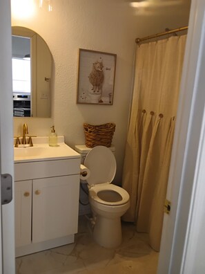 Ein Badezimmer mit Toilette, einem Waschbecken mit goldfarbener Armatur, einem Korbwäscheschrank auf dem Toilettendeckel, einem gerahmten Bild an der Wand und einem beige-farbenen Duschvorhang.