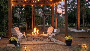 Una pérgola de madera con luces colgantes, un pozo de fuego y sillas Adirondack en un entorno boscoso.