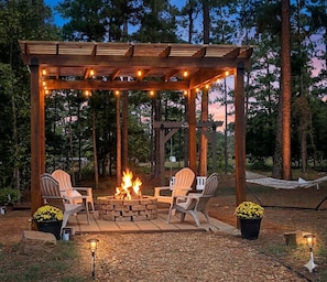 A wooden pergola with string lights, a fire pit, and Adirondack chairs in a wooded outdoor setting.