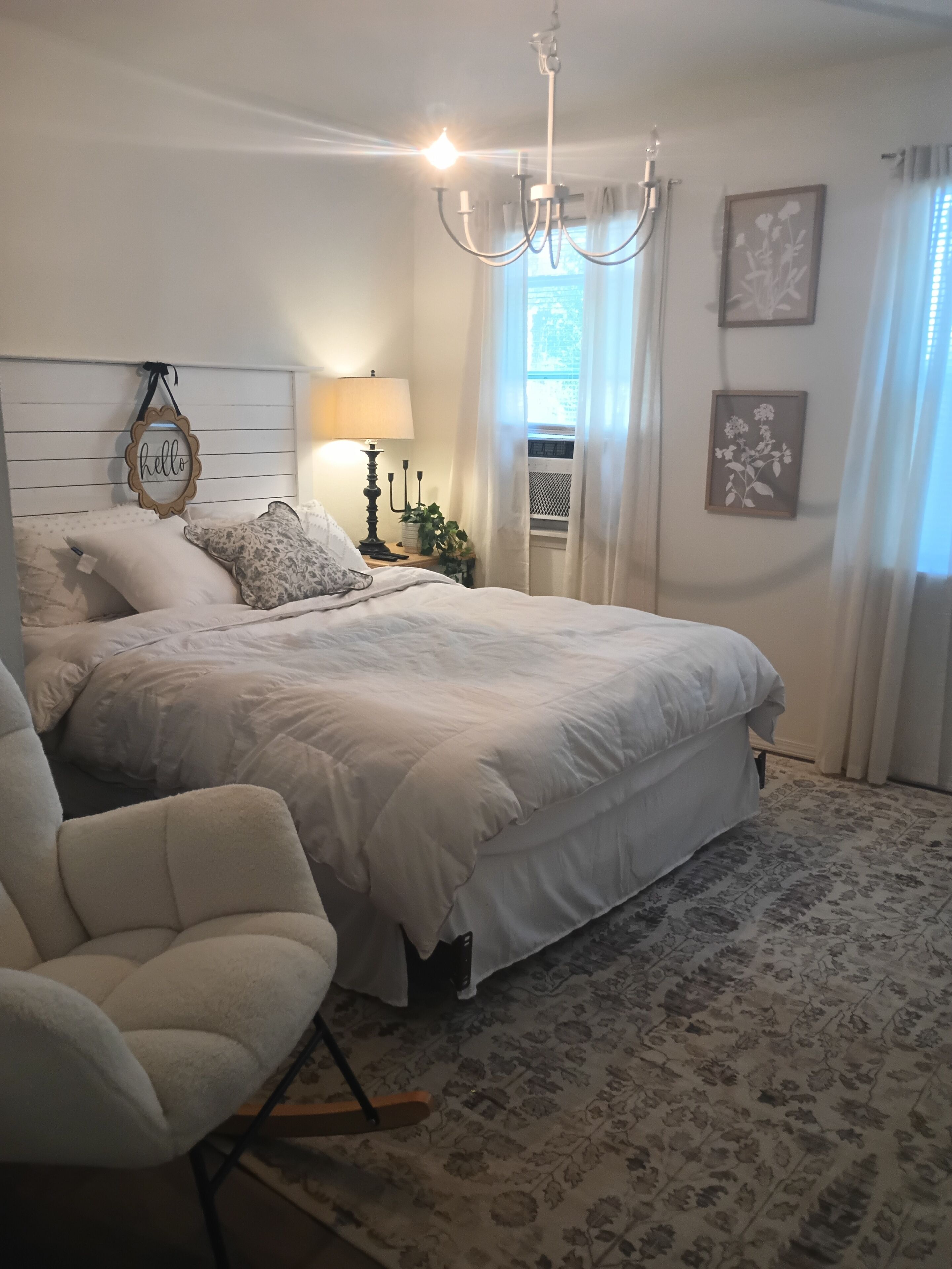A bedroom with a large bed, a bedside table with a lamp, a chair, and a chandelier.