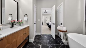Un baño moderno con piso de mármol negro, una bañera blanca y un mueble de tocador de madera.
