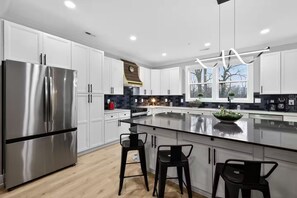 A modern kitchen with white cabinetry, a stainless steel refrigerator, a large island with bar stools, and a window providing natural light. - Beautiful 5-bedroom house in welcoming Fort Washington (Fort Washington)